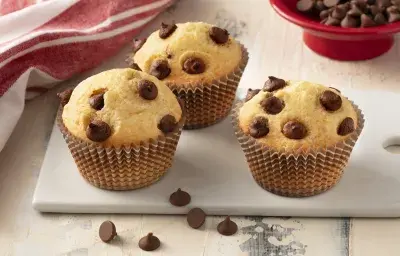 Fotografía en tonos de blanco y rojo de una encimera blanca con un banco, tres muffins encima. Al fondo un paño rojo y un recipiente redondo rojo con chispas de chocolate. Fotografía en tonos de blanco y rojo de una encimera blanca con un banco, tres muffins encima. Al fondo un paño rojo y un recipiente redondo rojo con chispas de chocolate.