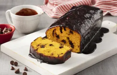 Fotografía en tonos blancos y rojos de una encimera blanca blanca con un plato rectangular blanco, sobre ella un pastel de zanahoria. Al fondo un paño de rayas rojas y blancas y un recipiente redondo blanco con salsa de chocolate. Fotografía en tonos blancos y rojos de una encimera blanca blanca con un plato rectangular blanco, sobre ella un pastel de zanahoria. Al fondo un paño de rayas rojas y blancas y un recipiente redondo blanco con salsa de chocolate.