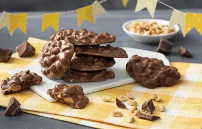 Fotografía en tonos amarillos con un paño a cuadros amarillo y trozos de mocoso chocolate de pie sobre una pizarra blanca. Banderas del partido Junina en cuadros amarillos y lunares en la parte superior y al fondo una pequeña olla blanca con cacahuetes. Fotografía en tonos amarillos con un paño a cuadros amarillo y trozos de mocoso chocolate de pie sobre una pizarra blanca. Banderas del partido Junina en cuadros amarillos y lunares en la parte superior y al fondo una pequeña olla blanca con cacahuetes.