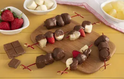 Fotografía en tonos amarillos en un banco de madera amarillo con una tabla de madera en el centro, con varias brochetas de frutas cubiertas de chocolate endurecido. A un lado, pequeñas ollas blancas con frutas: fresa, plátano y piña.