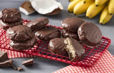 Fotografía en tonos rojos sobre una encimera gris con un respaldo rojo con las galletas de avena, cacao y coco. Al fondo, un racimo de plátanos y trozos de coco.