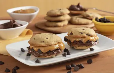 Fotografía en tonos amarillos en un banco de madera con un recipiente blanco en el centro con dos galletas rellenas de dulce de leche y crema de avellanas. Al fondo, ollas con chispas de chocolate, dulce de leche y una tabla con más galletas.