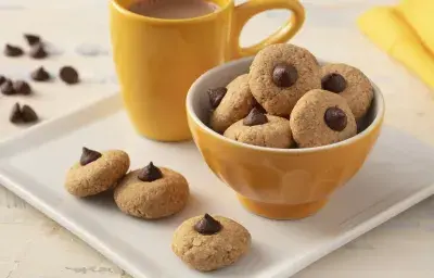 Fotografía en tonos de blanco y amarillo de una encimera blanca con un plato cuadrado, encima una taza amarilla, un recipiente amarillo redondo con galletas y tres galletas. Al fondo un paño amarillo y gotas de chocolate.