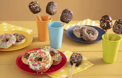 Fotografía en tonos amarillos sobre una encimera de madera amarilla con tres vasos de colores, azul, naranja y verde con piruletas de chocolate en su interior. En el centro, un plato rojo con tres donas cubiertas con chispitas de colores. Fotografía en tonos amarillos sobre una encimera de madera amarilla con tres vasos de colores, azul, naranja y verde con piruletas de chocolate en su interior. En el centro, un plato rojo con tres donas cubiertas con chispitas de colores.