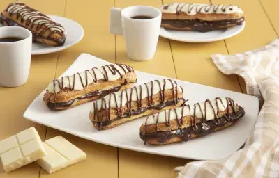 Fotografía en tonos amarillos en un banco de madera con un paño a cuadros color canela, un recipiente rectangular blanco en el centro con tres bombas de chocolate cubiertas de chocolate blanco. Al fondo, dos tazas de café y dos platos con dos bombas. Fotografía en tonos amarillos en un banco de madera con un paño a cuadros color canela, un recipiente rectangular blanco en el centro con tres bombas de chocolate cubiertas de chocolate blanco. Al fondo, dos tazas de café y dos platos con dos bombas.