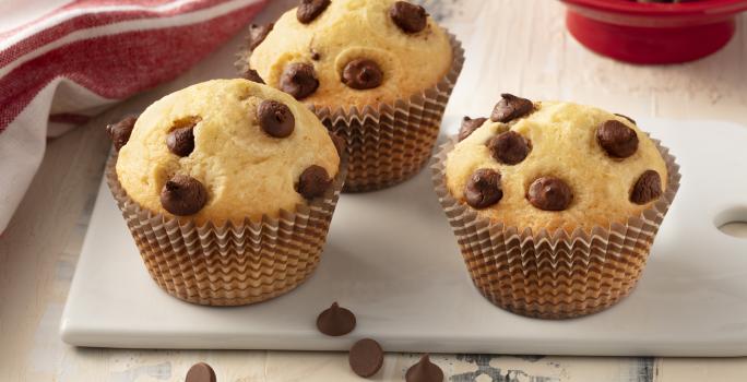 Fotografía en tonos de blanco y rojo de una encimera blanca con un banco, tres muffins encima. Al fondo un paño rojo y un recipiente redondo rojo con chispas de chocolate.