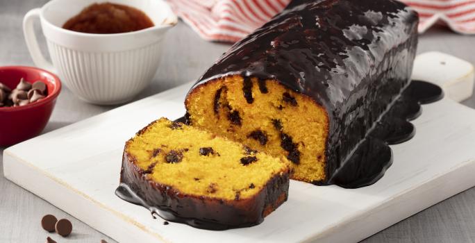 Fotografía en tonos blancos y rojos de una encimera blanca blanca con un plato rectangular blanco, sobre ella un pastel de zanahoria. Al fondo un paño de rayas rojas y blancas y un recipiente redondo blanco con salsa de chocolate.