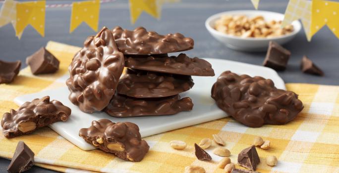 Fotografía en tonos amarillos con un paño a cuadros amarillo y trozos de mocoso chocolate de pie sobre una pizarra blanca. Banderas del partido Junina en cuadros amarillos y lunares en la parte superior y al fondo una pequeña olla blanca con cacahuetes.