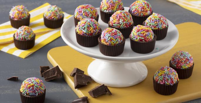 Fotografía en tonos de amarillo sobre una encimera gris oscuro con servilletas amarillas a rayas. En el centro, un tablero amarillo con un soporte de dulces blanco que sostiene los moldes de chocolate con brigadeiros con dulces de colores.