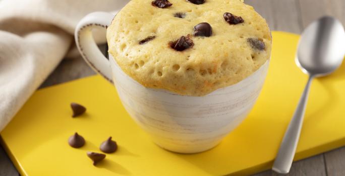 Fotografía en tonos marrones. blanco y amarillo de un banco de madera con un tablero amarillo, sobre él una taza blanca con pastel de taza y una cuchara. Al fondo una tela blanca.