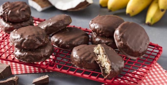 Fotografía en tonos rojos sobre una encimera gris con un respaldo rojo con las galletas de avena, cacao y coco. Al fondo, un racimo de plátanos y trozos de coco.