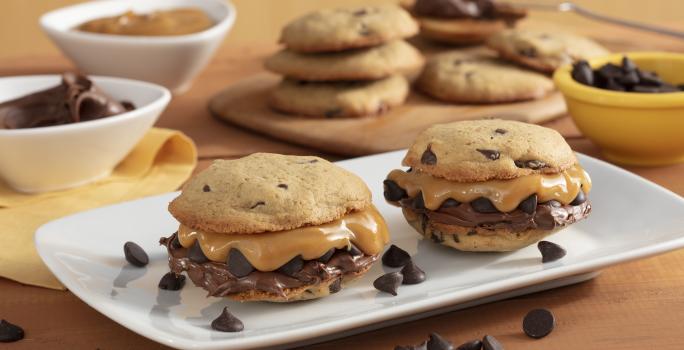 Fotografía en tonos amarillos en un banco de madera con un recipiente blanco en el centro con dos galletas rellenas de dulce de leche y crema de avellanas. Al fondo, ollas con chispas de chocolate, dulce de leche y una tabla con más galletas.