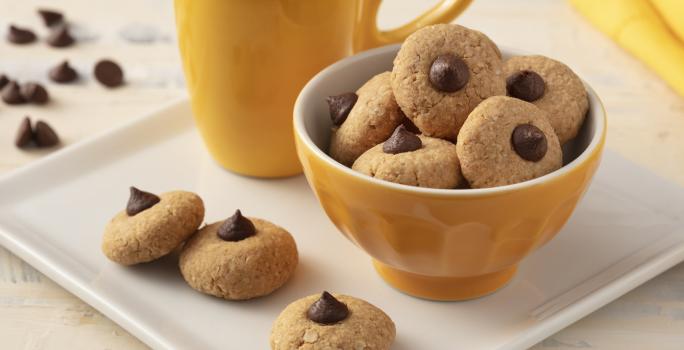 Fotografía en tonos de blanco y amarillo de una encimera blanca con un plato cuadrado, encima una taza amarilla, un recipiente amarillo redondo con galletas y tres galletas. Al fondo un paño amarillo y gotas de chocolate.