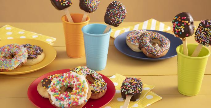 Fotografía en tonos amarillos sobre una encimera de madera amarilla con tres vasos de colores, azul, naranja y verde con piruletas de chocolate en su interior. En el centro, un plato rojo con tres donas cubiertas con chispitas de colores.