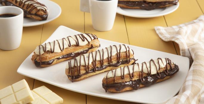 Fotografía en tonos amarillos en un banco de madera con un paño a cuadros color canela, un recipiente rectangular blanco en el centro con tres bombas de chocolate cubiertas de chocolate blanco. Al fondo, dos tazas de café y dos platos con dos bombas.