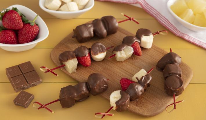 Fotografía en tonos amarillos en un banco de madera amarillo con una tabla de madera en el centro, con varias brochetas de frutas cubiertas de chocolate endurecido. A un lado, pequeñas ollas blancas con frutas: fresa, plátano y piña.