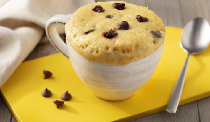 Fotografía en tonos marrones. blanco y amarillo de un banco de madera con un tablero amarillo, sobre él una taza blanca con pastel de taza y una cuchara. Al fondo una tela blanca. Fotografía en tonos marrones. blanco y amarillo de un banco de madera con un tablero amarillo, sobre él una taza blanca con pastel de taza y una cuchara. Al fondo una tela blanca.