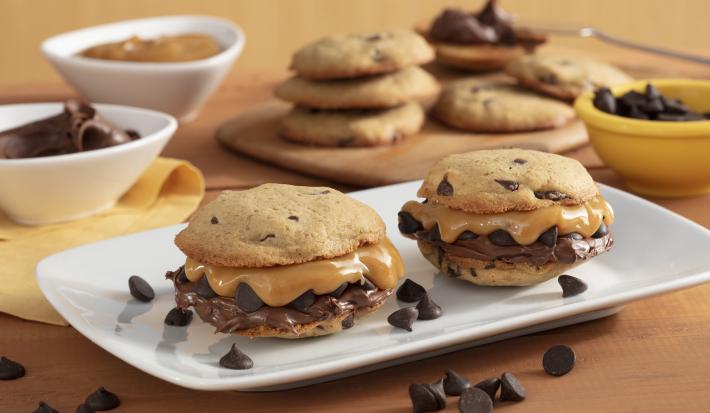 Fotografía en tonos amarillos en un banco de madera con un recipiente blanco en el centro con dos galletas rellenas de dulce de leche y crema de avellanas. Al fondo, ollas con chispas de chocolate, dulce de leche y una tabla con más galletas.