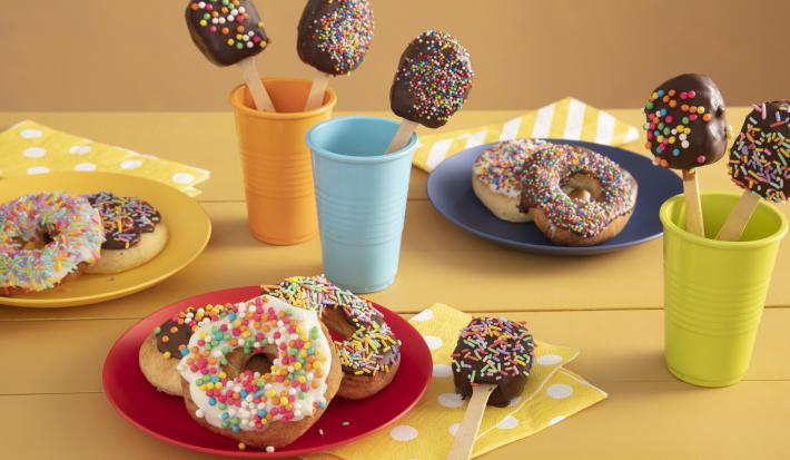 Fotografía en tonos amarillos sobre una encimera de madera amarilla con tres vasos de colores, azul, naranja y verde con piruletas de chocolate en su interior. En el centro, un plato rojo con tres donas cubiertas con chispitas de colores. Fotografía en tonos amarillos sobre una encimera de madera amarilla con tres vasos de colores, azul, naranja y verde con piruletas de chocolate en su interior. En el centro, un plato rojo con tres donas cubiertas con chispitas de colores.
